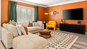 A living room with a beige sofa, a yellow armchair, a coffee table, a TV, and a wall-mounted shelf. - Epic Desert Stay Pool Hot Tub Sports Court (Scottsdale)