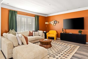 Ein Wohnzimmer mit einem beigefarbenen Sofa, einem gelben Sessel, einem Couchtisch, einem Fernseher und einem Wandregal.