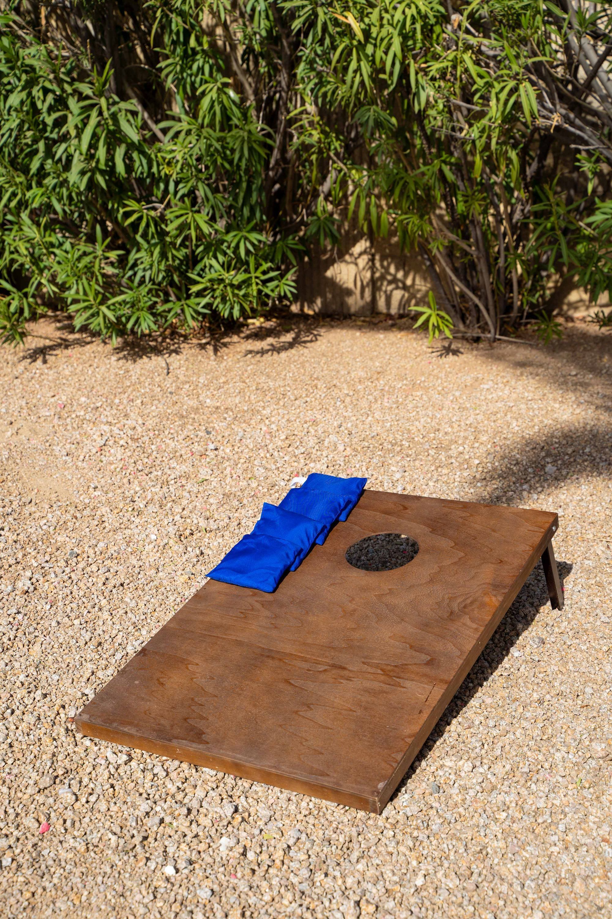 Une planche carrée en bois recouverte d’un tissu bleu et percée d’un trou circulaire, posée sur du gravier, avec un buisson en arrière-plan.