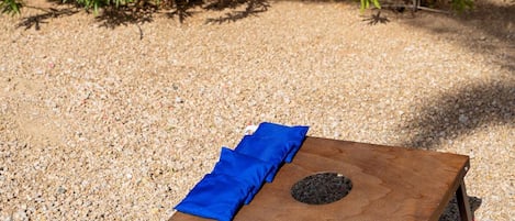 Une planche carrée en bois recouverte d’un tissu bleu et percée d’un trou circulaire, posée sur du gravier, avec un buisson en arrière-plan.