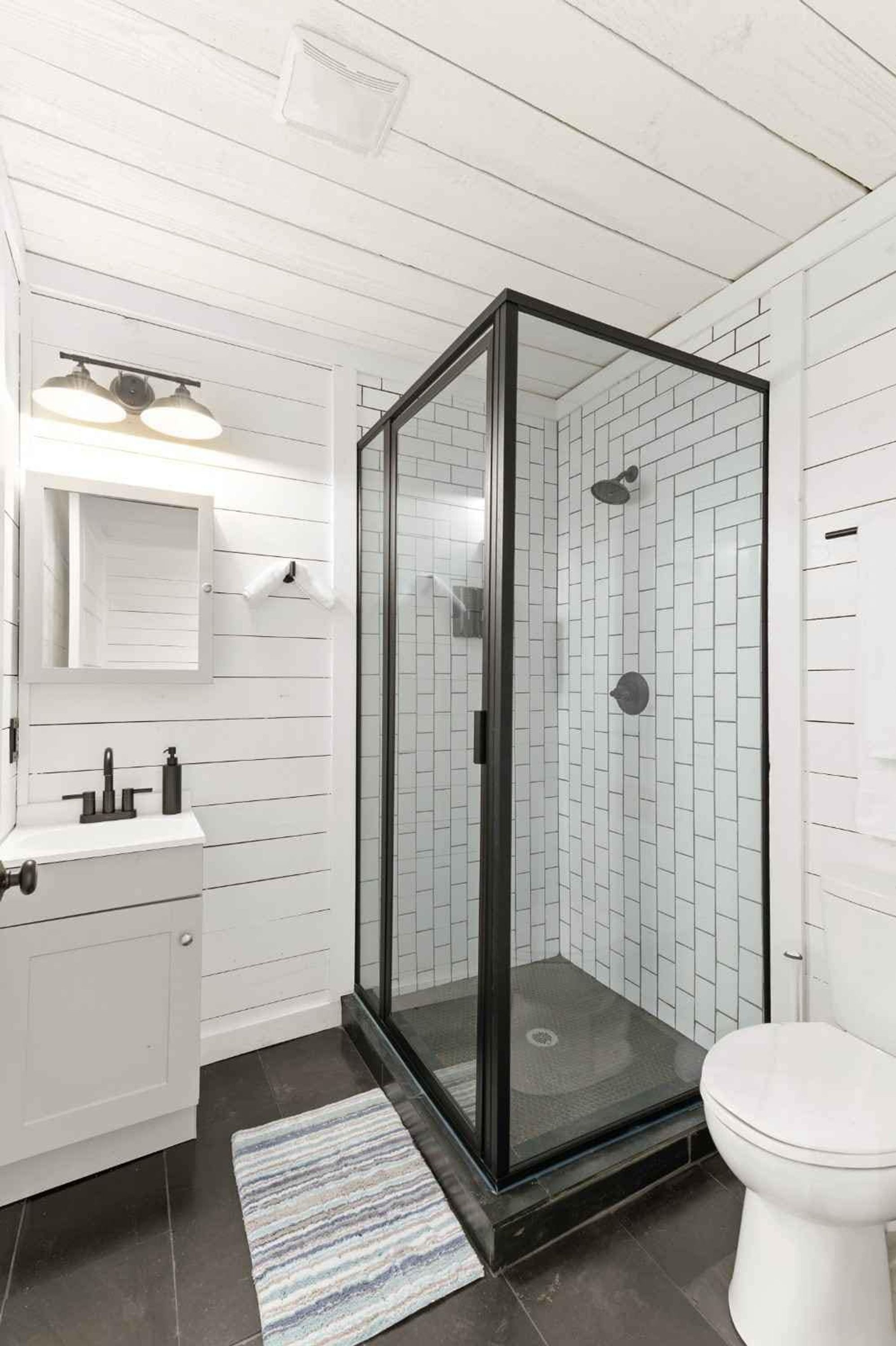 Un baño con ducha acristalada, paredes de azulejos blancos, lavamanos blanco y un inodoro.