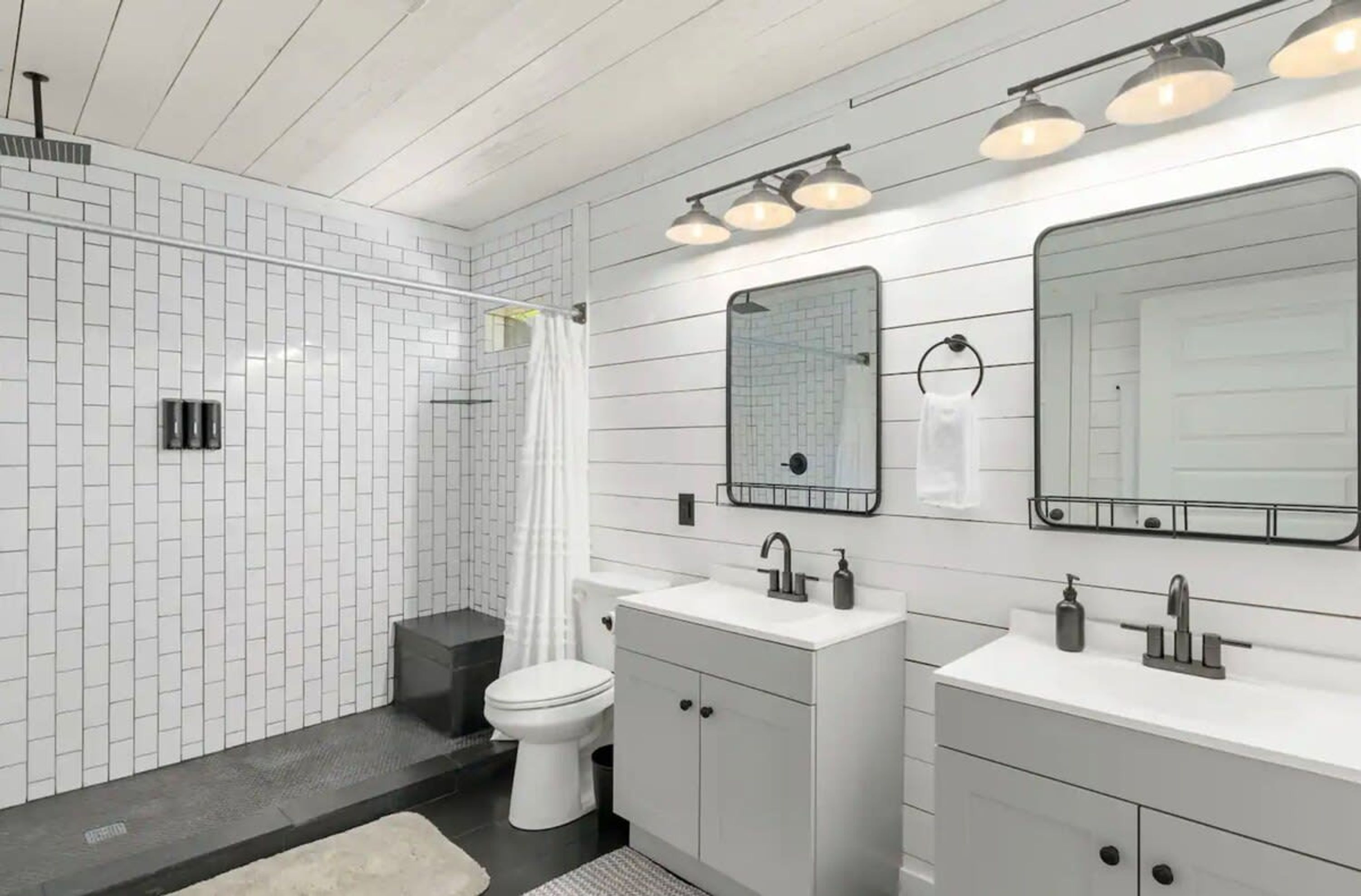 Un baño moderno con paredes de azulejos blancos, ducha, inodoro y dos lavamanos con espejos encima.