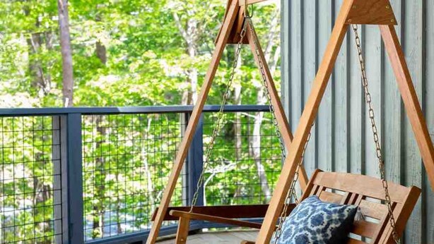 Een houten schommelstoel met kussen, opgehangen aan kettingen, op een houten terras met een reling, met uitzicht op een groen landschap.