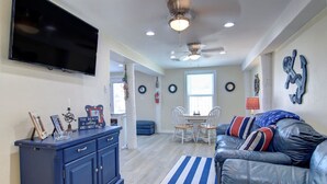 A living room with a blue leather couch, a flat-screen TV, a ceiling fan, and a dining area with a table and chairs. - Nauti Mermaid Private Pool Sleeps 14 Dog Friendly (Port Aransas)