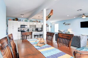 A spacious living area with a dining table, wooden chairs, a kitchen with white cabinets, and a TV mounted on the wall. - Nauti Mermaid Private Pool Sleeps 14 Dog Friendly (Port Aransas)