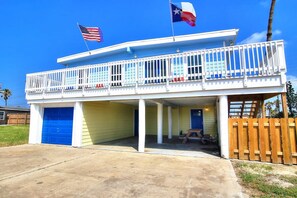 A two-story building with a balcony, a blue garage door, and a flagpole with two flags. - Nauti Mermaid Private Pool Sleeps 14 Dog Friendly (Port Aransas)