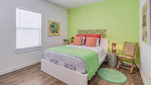 Un dormitorio con cama, ventana con persianas, pared verde, planta decorativa y una silla.