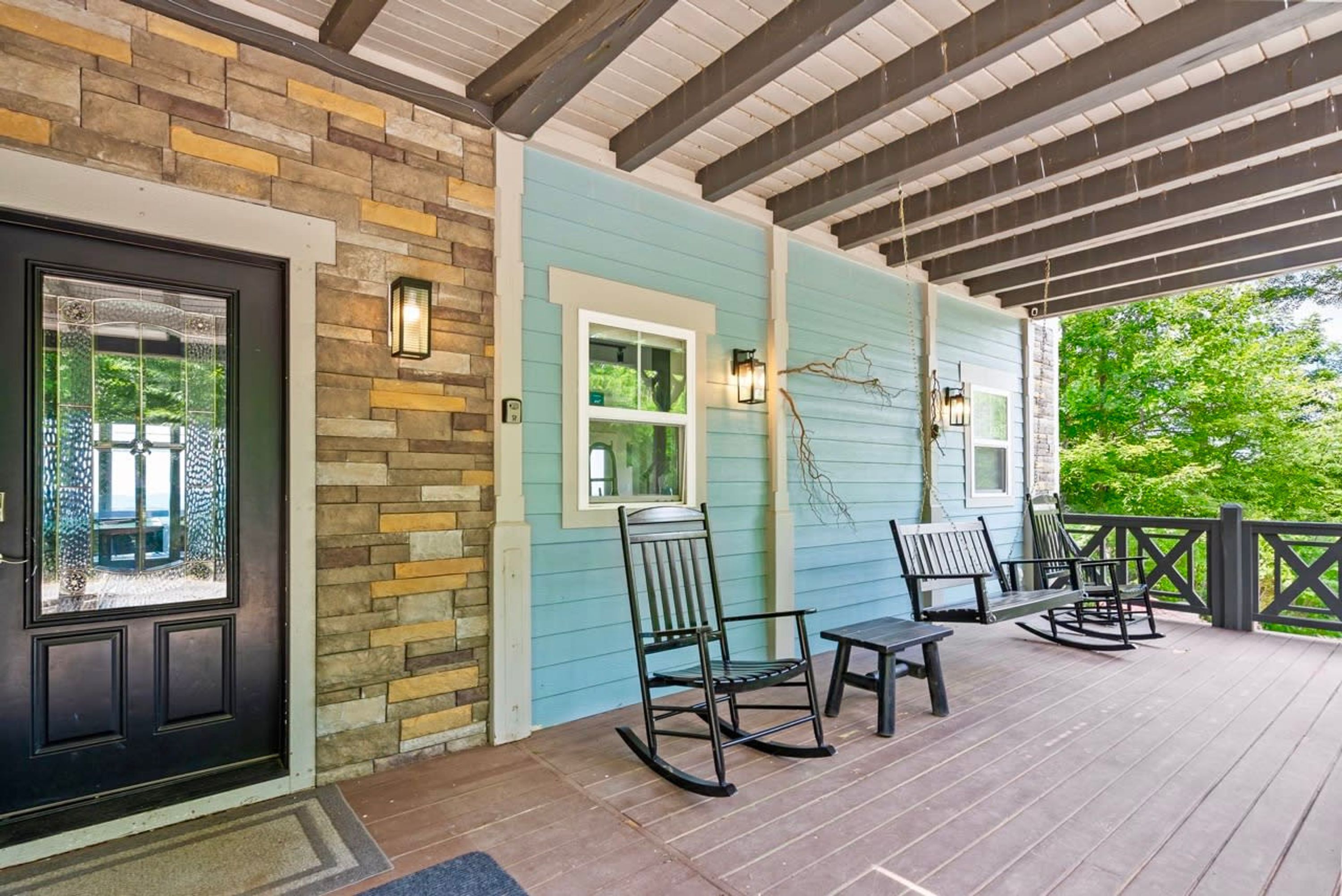 A covered porch with a stone wall, a black door, and a blue exterior wall. There are two rocking chairs and a table on the porch.