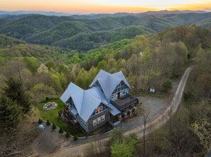A two-story cabin with a stone foundation and metal roof, surrounded by a forested landscape with a clear view of the surrounding hills. - 10min to Hatley Pointe Views Hottub Firepit (Mars Hill)
