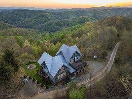 A two-story cabin with a stone foundation and metal roof, surrounded by a forested landscape with a clear view of the surrounding hills.