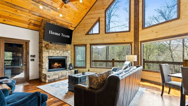 A spacious living area with a stone fireplace, a television mounted on the wall, and a large window overlooking a forested area. - Blue Ridge Cabin Stunning Views Hot Tub Game Room (Cherry Log)