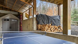 A ping pong table in a covered area with a stack of firewood and a door in the background. - Blue Ridge Cabin Stunning Views Hot Tub Game Room (Cherry Log)