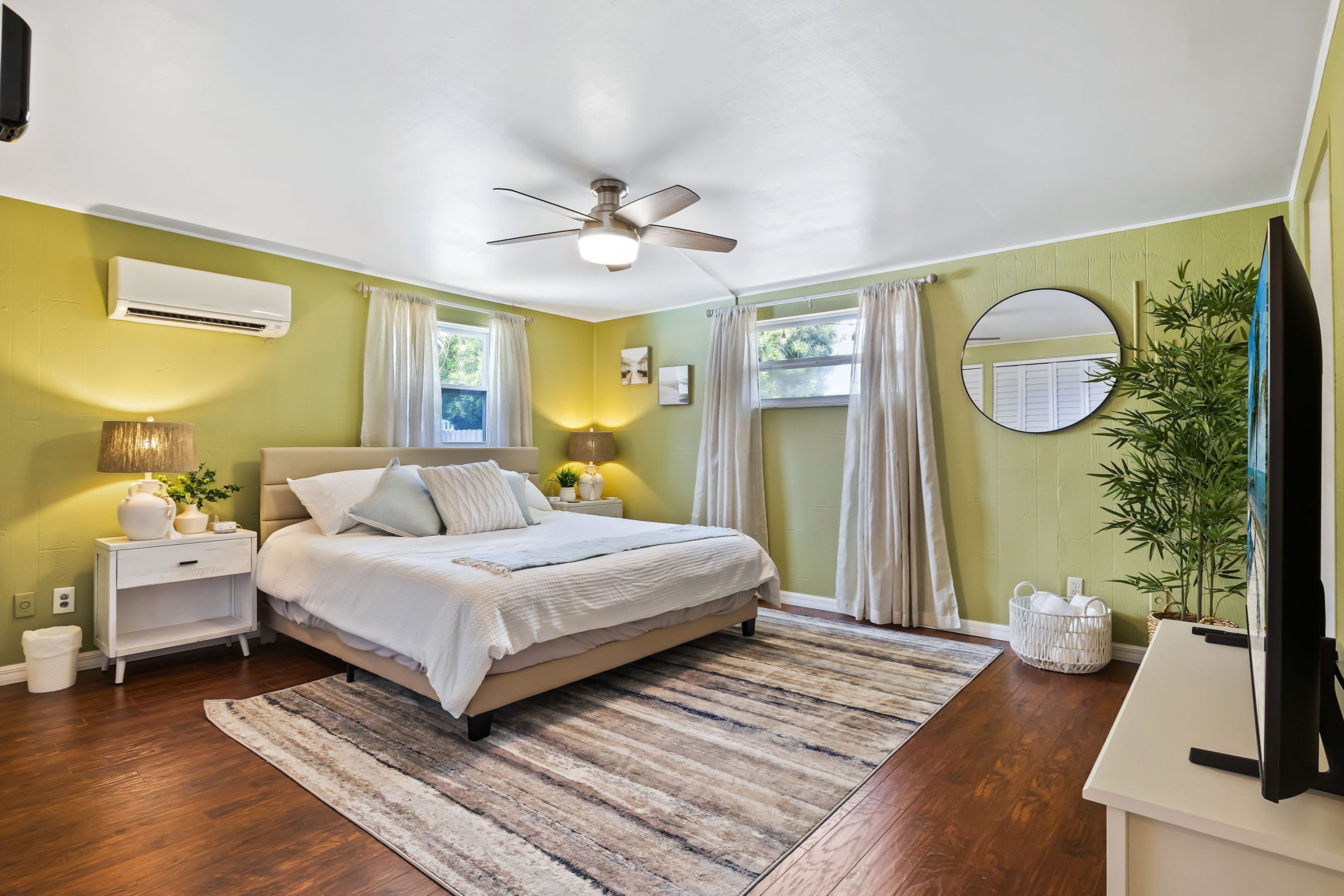 A bedroom with a bed, bedside tables, an air conditioner, a ceiling fan, a window with curtains, a mirror, and a plant.