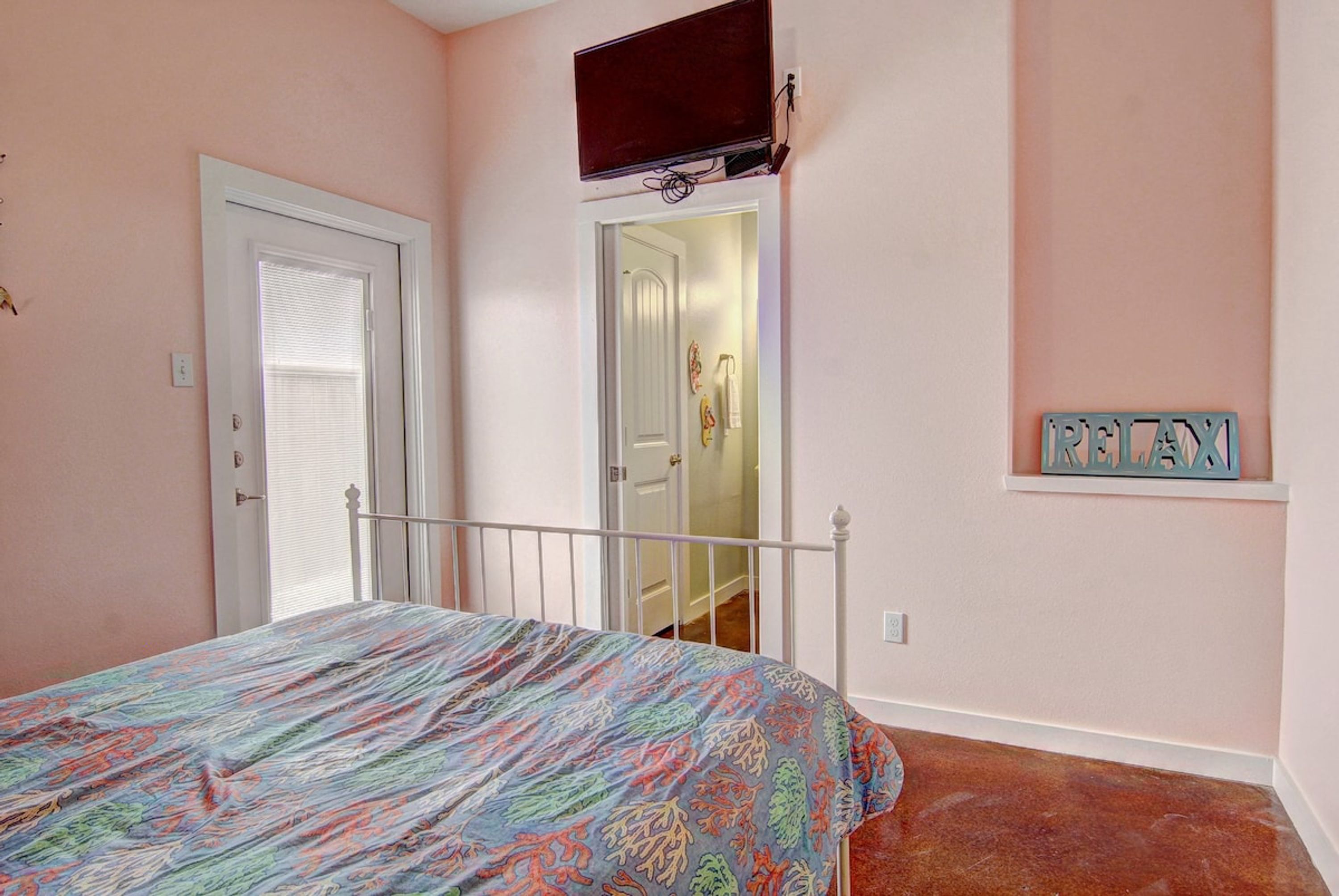 Ein Schlafzimmer mit einem Bett, einer Tür, einem an der Wand montierten Fernseher und einem kleinen Regal mit Namensschild.