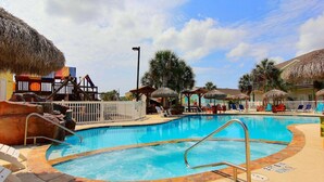 A swimming pool with lounge chairs and thatched umbrellas, surrounded by a fence and palm trees. - Gone Coastal Private Breezeway Family Friendly (Port Aransas)
