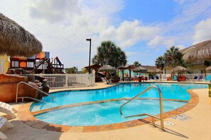 A swimming pool with lounge chairs and thatched umbrellas, surrounded by a fence and palm trees. - Gone Coastal Private Breezeway Family Friendly (Port Aransas)
