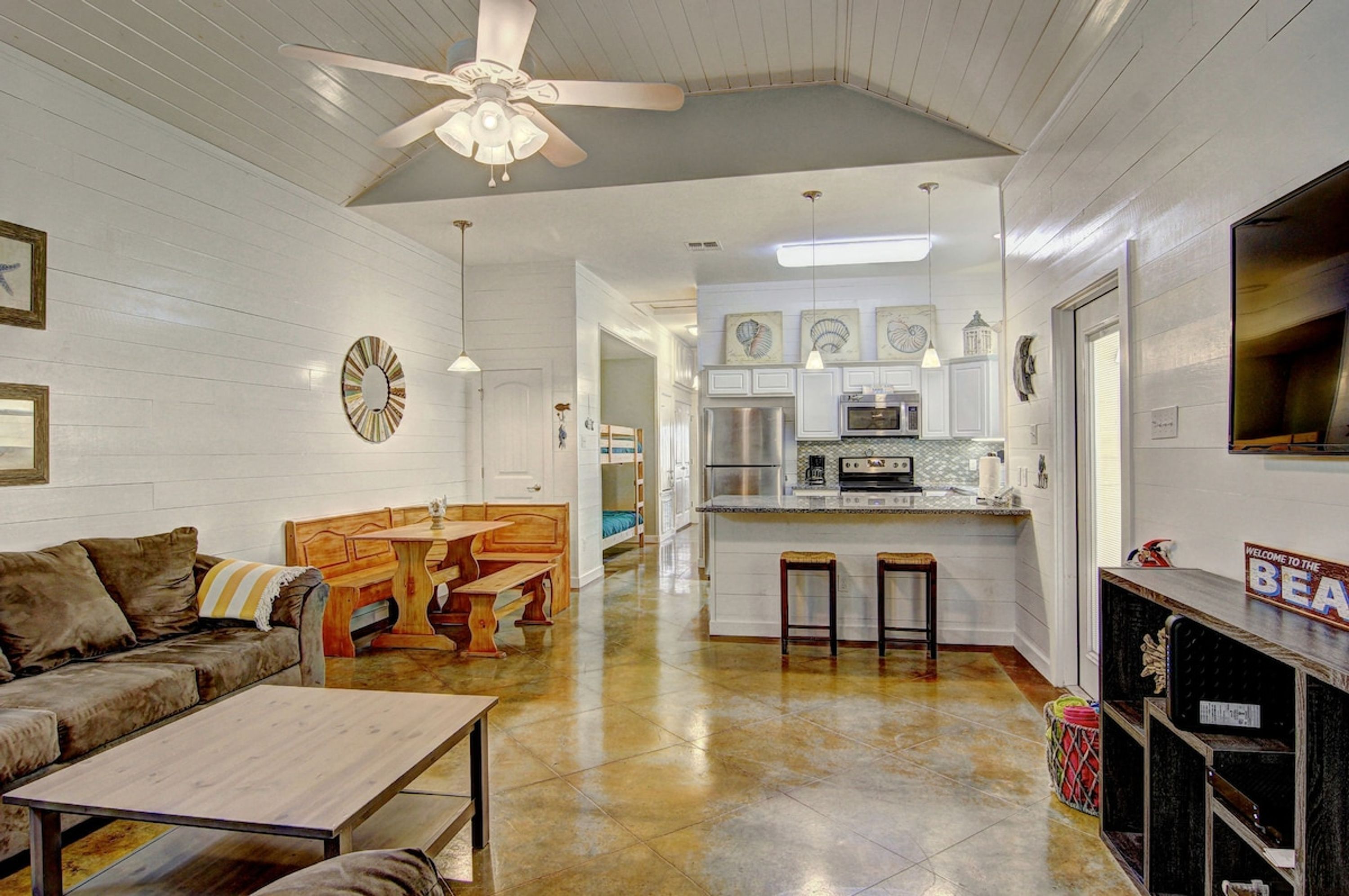 A spacious living area with a ceiling fan, a dining table, and a sofa. A kitchen with a central island and bar stools is visible in the background.