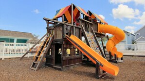 A wooden play structure with a slide and climbing elements, surrounded by a white fence. - Gone Coastal Private Breezeway Family Friendly (Port Aransas)