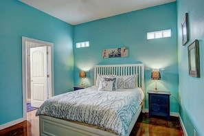 Un dormitorio con cama, dos mesitas de noche, una puerta y un cuadro en la pared.