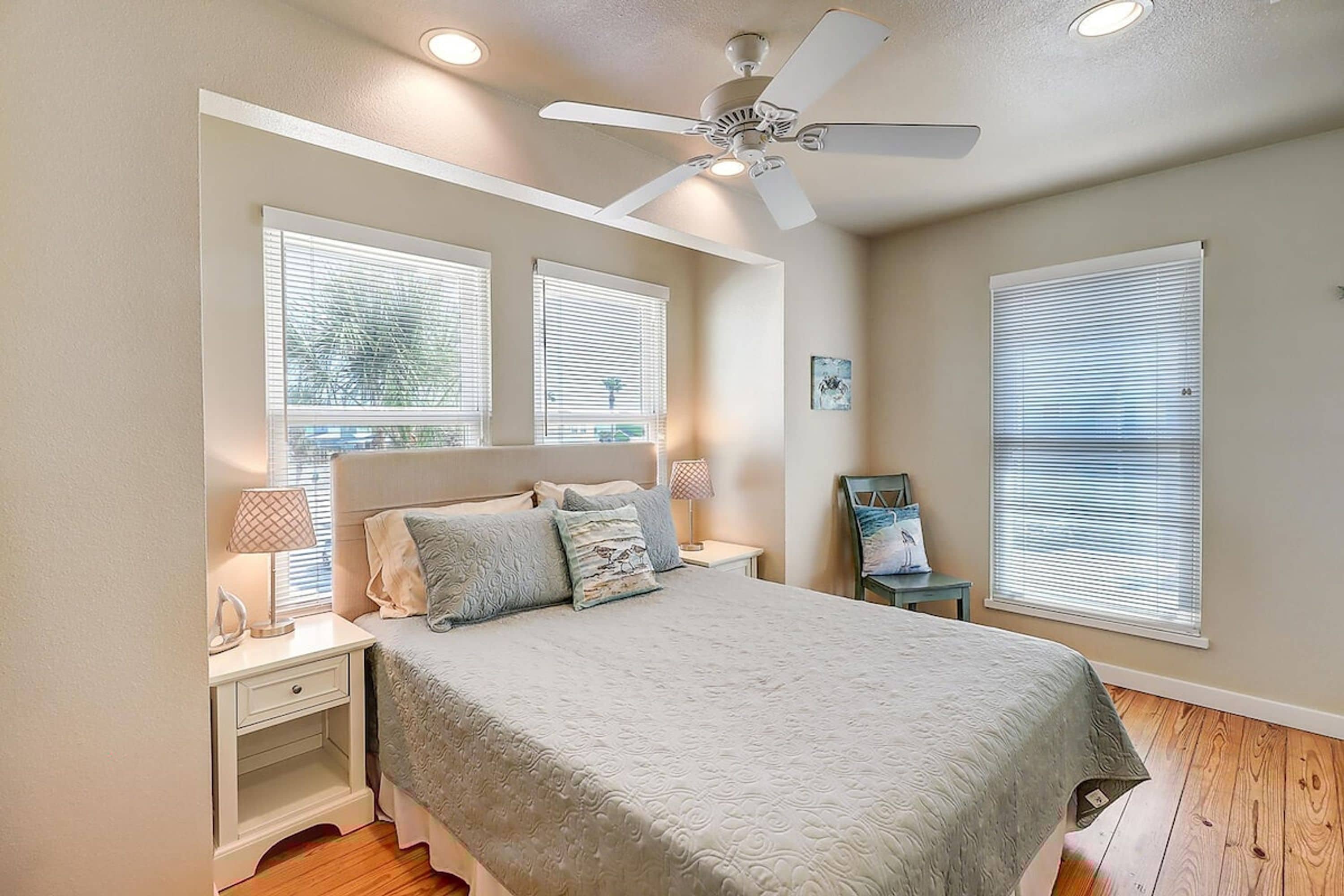 A bedroom with a bed, nightstands, a ceiling fan, and large windows with blinds.