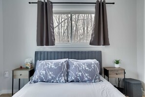 A bedroom with a bed, a bedside table, a nightstand, a window with curtains, and a view of trees outside.