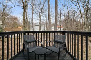 Une terrasse avec deux chaises et une petite table, entourée d’une rambarde en bois, donnant sur une zone boisée avec des maisons en arrière-plan.