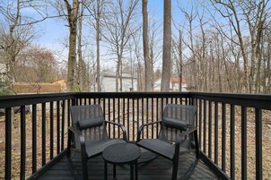 Terraza con dos sillas y una pequeña mesa, rodeada por baranda de madera, con vista a una zona boscosa y casas al fondo.
