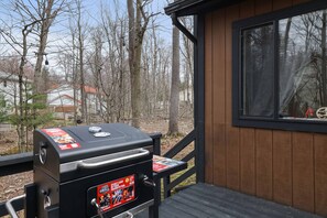 A barbecue grill with a fuel can on a wooden deck, surrounded by trees and a cabin.