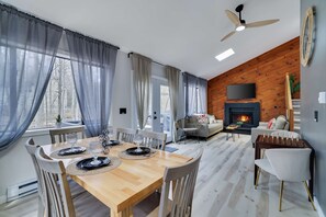 A modern living room with a fireplace, a dining area with a wooden table and chairs, and a ceiling fan. - 3BR Home w Outdoor Fun Lake Fire Pit Games Grill (Tobyhanna)