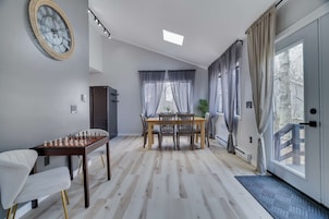 A dining area with a chess table, a dining table with chairs, a large window with curtains, and a door with a mat.