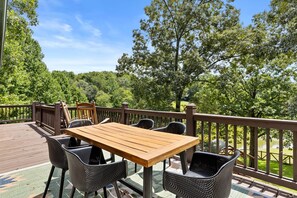 A wooden deck with a table and chairs, surrounded by a wooden railing, overlooking a forested area. - Lake Views Hot Tub Fire Pit Micro Events (Morganton)
