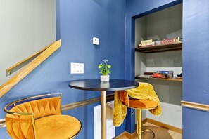 A room with a blue wall, a round table with two chairs, a small plant on the table, a thermostat on the wall, and a shelf with books and a puzzle.