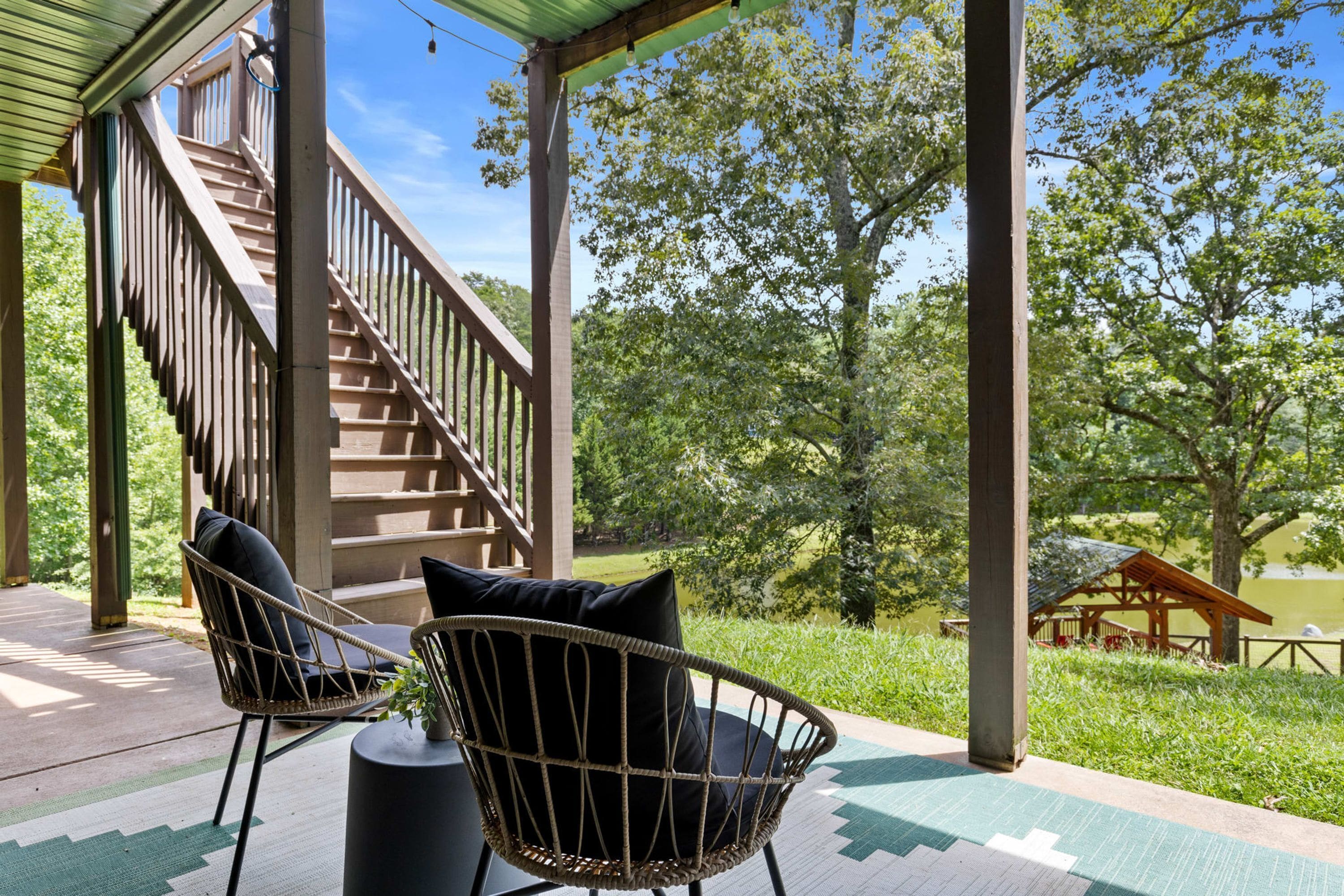 Un espace extérieur abrité, doté d’un escalier en bois, de deux fauteuils en osier avec des coussins, et offrant une vue sur un lac et des arbres.