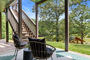 A covered outdoor area with a wooden staircase, two wicker chairs with cushions, and a view of a lake and trees.