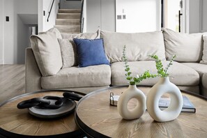 Ein modernes Wohnzimmer mit einem runden Holztisch, zwei weiße Vasen mit Pflanzen und einem Sofa mit beigefarbenen Kissen und einem blauen Kopfkissen.