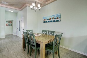 A dining room with a wooden table and chairs, a chandelier, and a painting of seashells on the wall. - Piggy s Perch- 2 Balconies Direct Pool Access (Port Aransas)