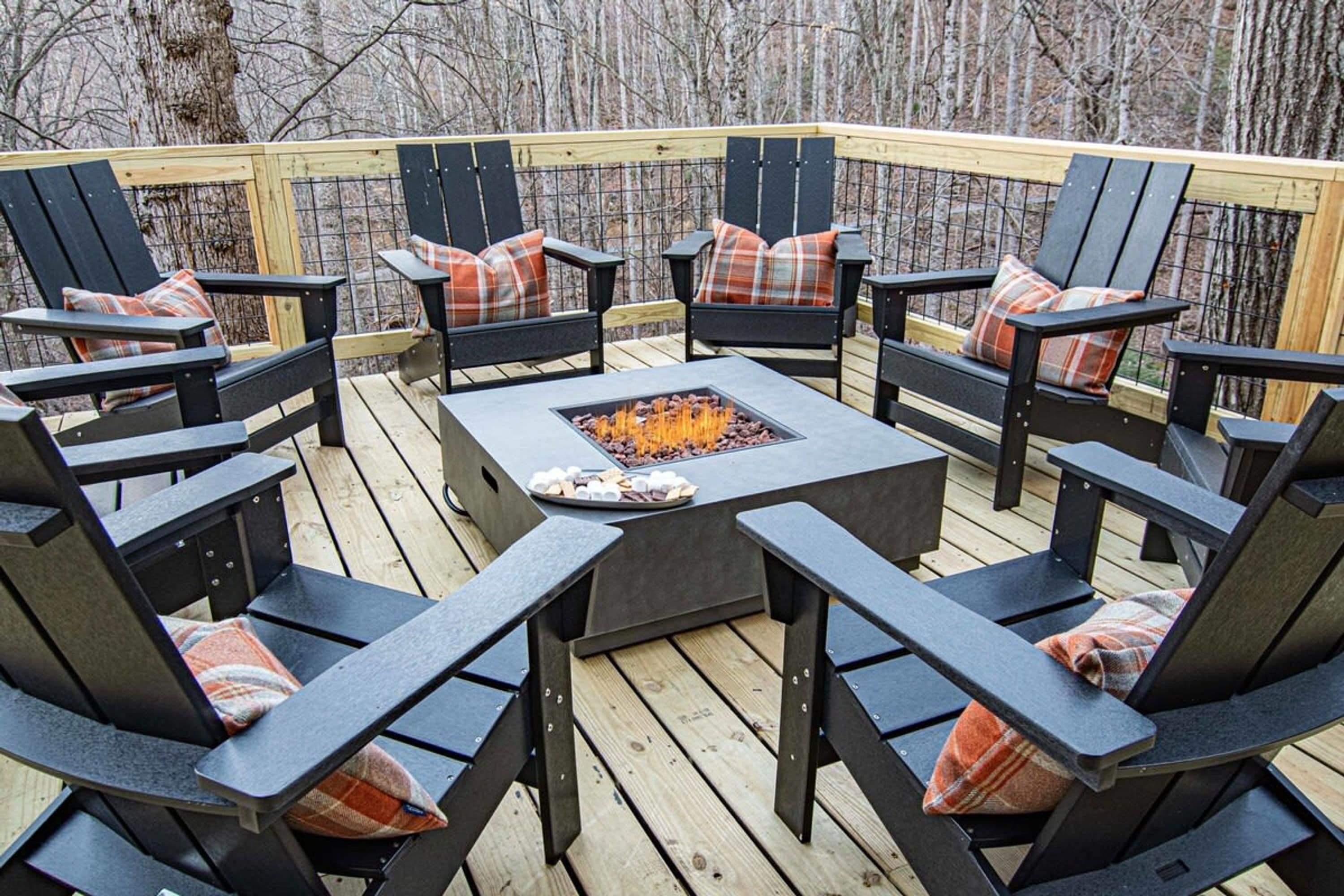 Une terrasse extérieure avec des fauteuils Adirondack noirs et des coussins à carreaux, un foyer carré au centre, entouré d’une terrasse en bois et d’une rambarde en métal.