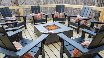 An outdoor deck with black Adirondack chairs and plaid cushions, a square fire pit in the center, surrounded by a wooden deck and a metal railing.