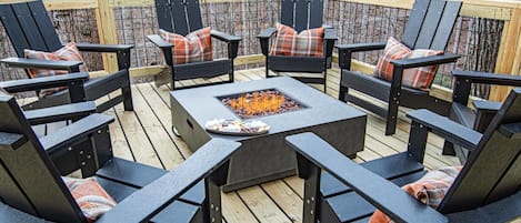Une terrasse extérieure avec des fauteuils Adirondack noirs et des coussins à carreaux, un foyer carré au centre, entouré d’une terrasse en bois et d’une rambarde en métal.