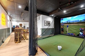 Ein Raum mit einer Golf-Simulationsanlage, einschließlich eines Bildschirms, der ein Golfspiel zeigt, einem mit Netzen versehenen Bereich zum Üben von Schwüngen und einer Bar mit Hockern.