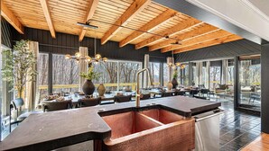 A modern kitchen with a large copper sink, wooden ceiling beams, and large windows offering views of trees. - 7k sq ft Mountain Estate -the Most Epic Experience (Asheville)