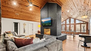 A spacious living area with a large fireplace, a comfortable sofa, and a television mounted on a stone wall. - 7k sq ft Mountain Estate -the Most Epic Experience (Asheville)