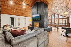A spacious living area with a large fireplace, a comfortable sofa, and a television mounted on a stone wall. - 7k sq ft Mountain Estate -the Most Epic Experience (Asheville)