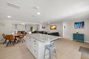 A modern kitchen with a central island, dining area with chairs, a sofa, and a TV mounted on the wall. - 3 Miles to Honeymoon Island Firepit Pool (Palm Harbor)
