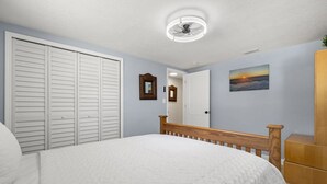 Um quarto com cama, guarda-roupa, espelho, quadro na parede e ventoinha no teto.