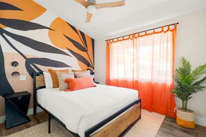 Een slaapkamer met een bed, een plafondventilator, een nachtkastje, een wandlamp, een raam met oranje gordijnen en een kamerplant.