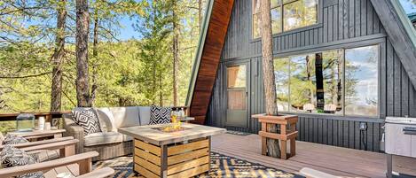Una terraza de madera moderna con mobiliario exterior, un hogar para fuego y vistas al bosque circundante.