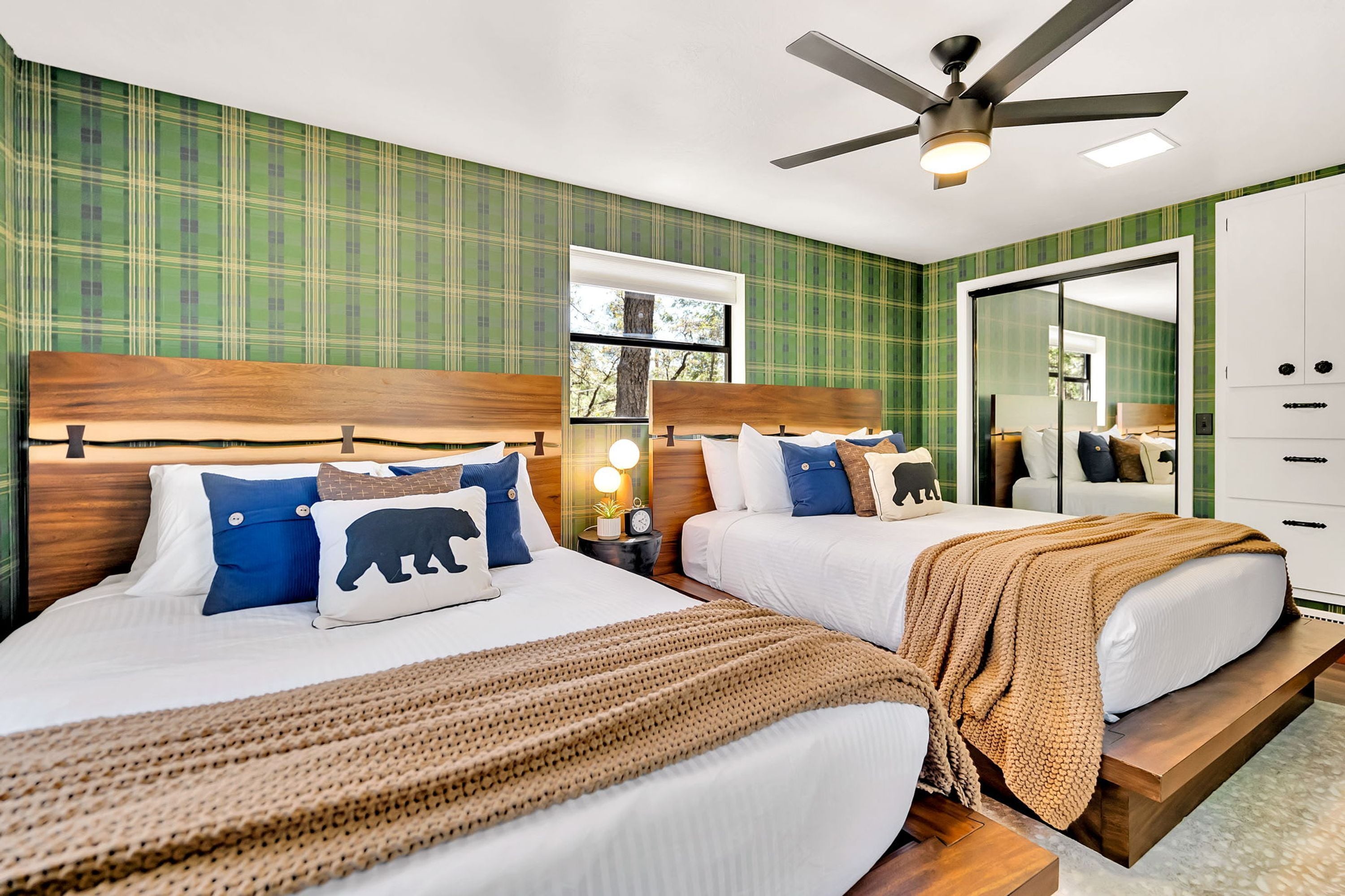 A hotel room with two beds, a ceiling fan, a large mirror, and a window with a view of trees.