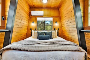 Un dormitorio acogedor con techo de madera, una cama grande con muchas almohadas y una ventana con vistas a árboles.