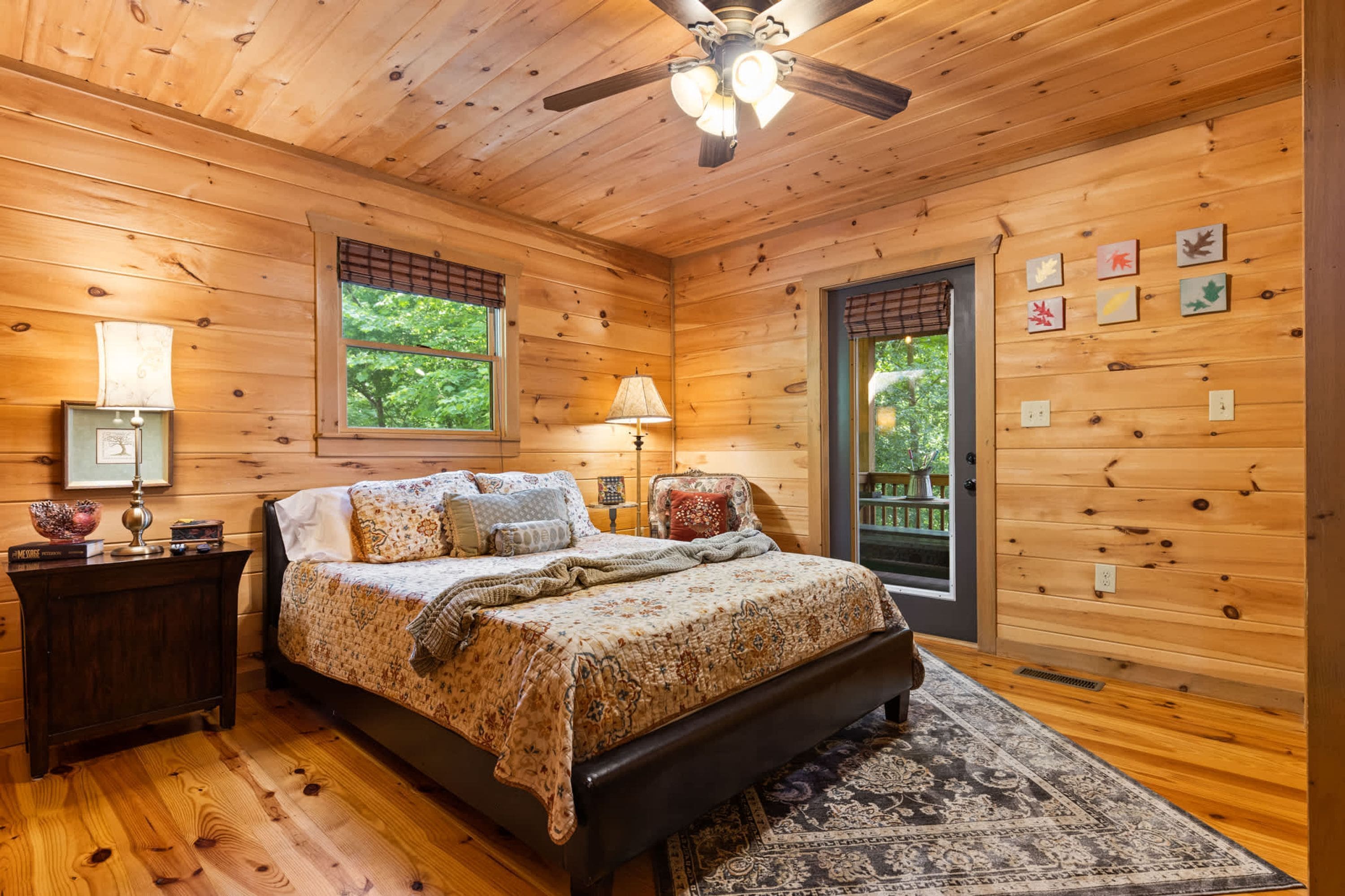 A wooden cabin bedroom with a bed, nightstands, a ceiling fan, and a view of the outdoors.